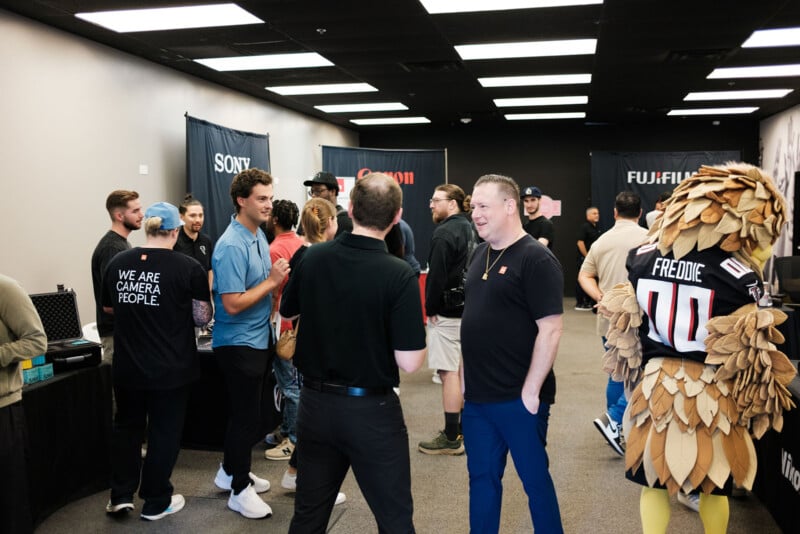 A group of people chat and mingle at an indoor event with camera brand banners on the walls. A person in a bird mascot costume labeled "FREDDIE 01" stands to the right. The atmosphere is casual and social.