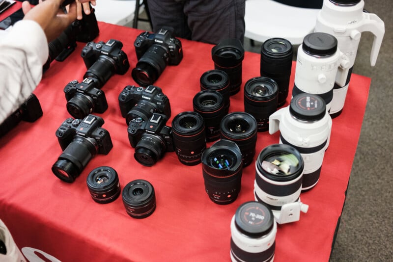 A red tablecloth displays several black DSLR cameras, various camera lenses, and a few large white telephoto lenses. Two people are partially visible, reaching toward the equipment.
