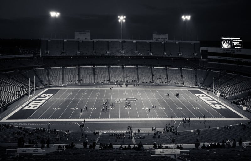 A black and white photo of an empty football stadium at night, with stadium lights illuminating the field. The word "BILLS" is visible in each end zone, and only a few people are scattered in the stands and on the field.