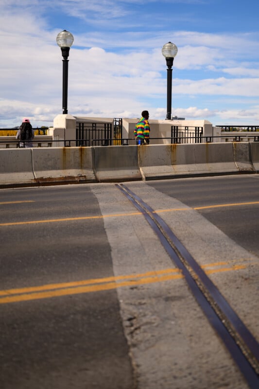 Un hombre con un colorido suéter a rayas estaba junto a la cerca entre dos luces de la calle en el puente, mientras que el otro caminaba hacia la izquierda. El cielo está nublado y las pistas en primer plano cruzan el camino.