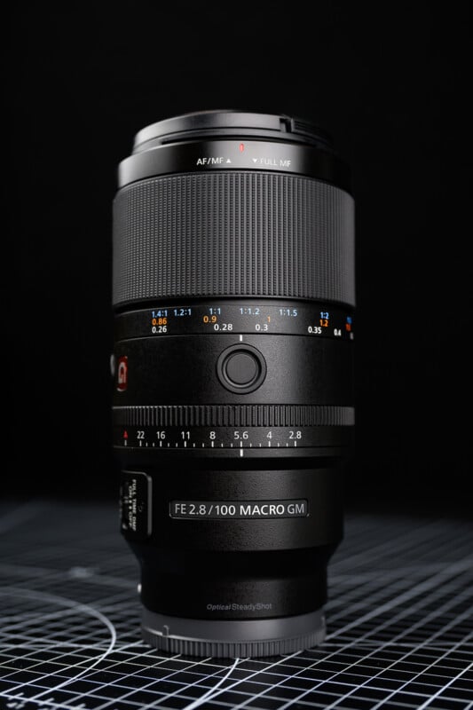A close-up photo of a Sony FE 100mm f/2.8 GM macro camera lens standing upright on a grid-patterned surface, with a black background. The lens details, controls, and markings are clearly visible.