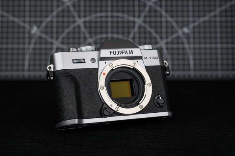 Cuerpo de cámara sin espejo Fujifilm X-T30 sin lente colocada, colocado sobre una superficie negra con fondo de cuadrícula geométrica.