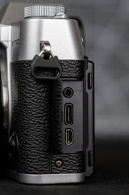 Primer plano del costado de una cámara digital plateada y negra, que muestra tres puertos abiertos: conector para auriculares, puerto HDMI y puerto USB, con la cubierta de goma del puerto doblada hacia atrás.