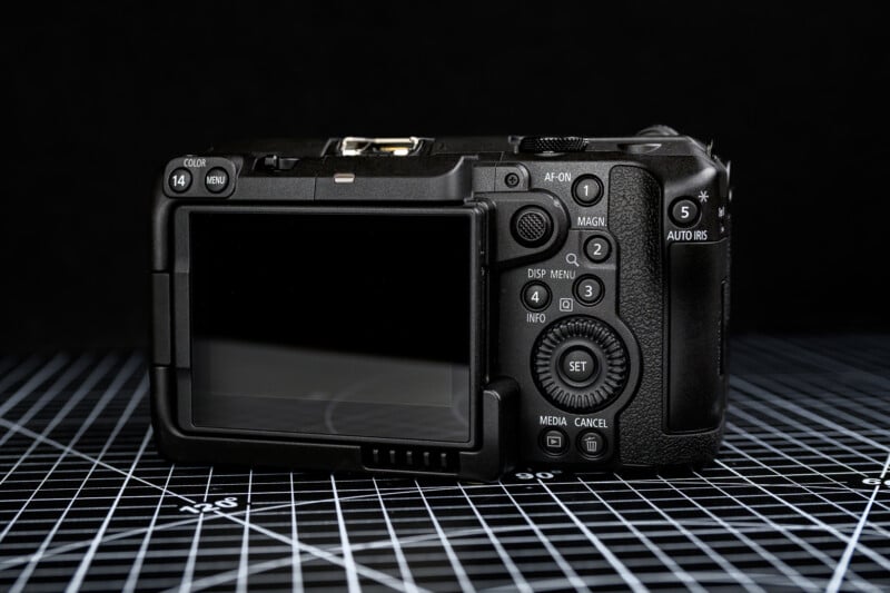 A digital camera shown from the back, displaying its screen and various buttons. The camera is placed on a black grid-patterned surface, with a dark background behind it.