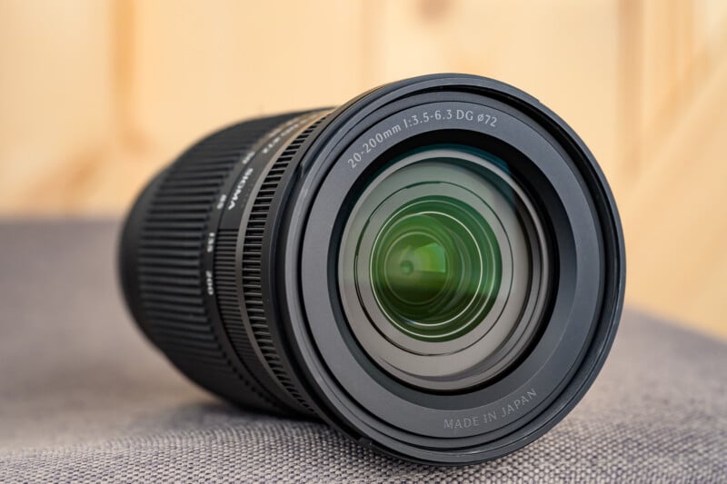 A close-up view of a camera lens lying on its side, showing the front glass element and detailed text around the rim indicating "20-200mm 1:3.5-6.3 DG Ø72" and "Made in Japan.