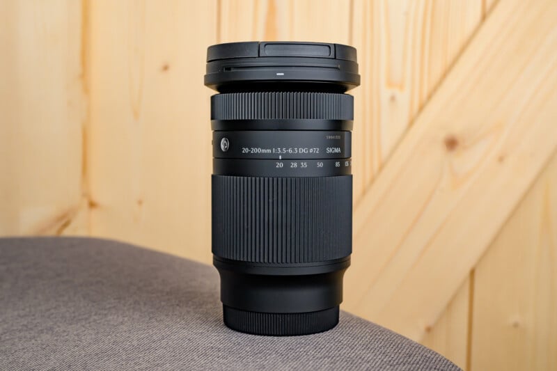 A Sigma 20-200mm camera lens stands upright on a gray fabric surface with a light wooden wall in the background. The lens is black with textured grip rings and clear white markings.