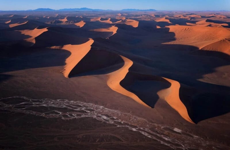 Aerial view of tall, orange sand dunes casting long shadows on a dark desert landscape, with a dry riverbed winding through the foreground and distant mountains on the horizon.