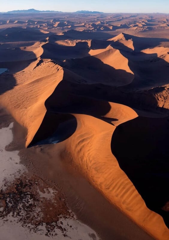 Aerial view of large, orange sand dunes casting dramatic shadows across the desert, with distant mountains under a clear sky.