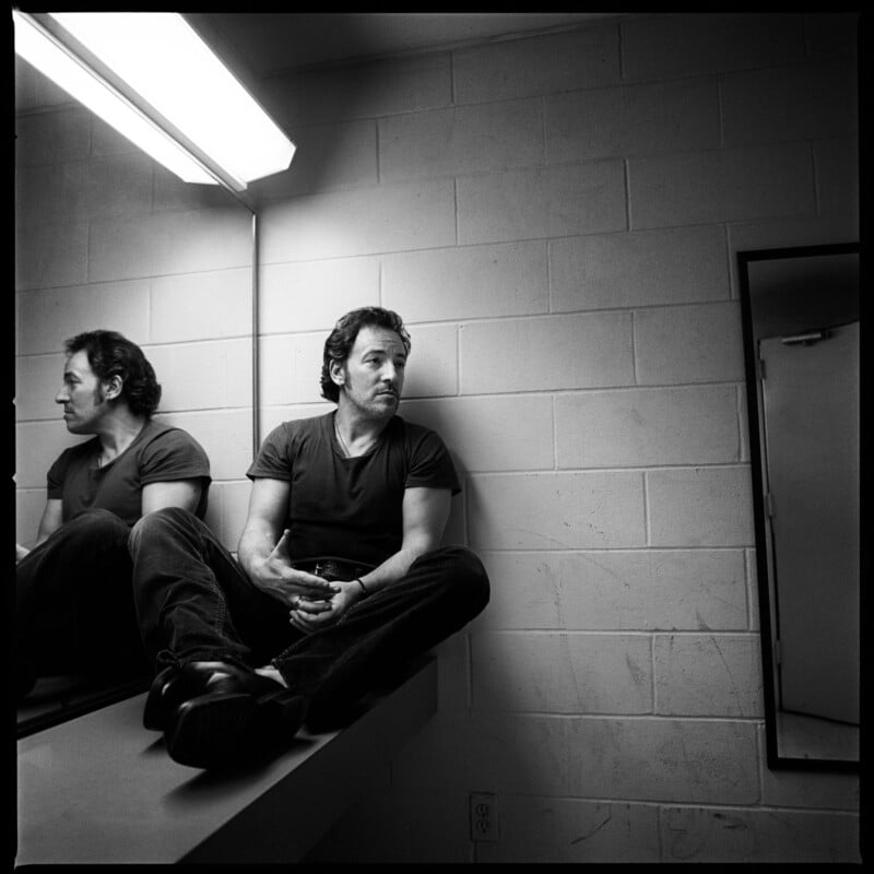A man in a dark t-shirt and jeans sits cross-legged on a counter in a stark room, looking to the side, with his reflection visible in the mirror beside him. The setting appears to be a backstage or dressing room.