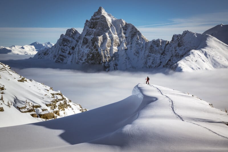 A lone skier stands atop a snowy ridge, overlooking mist-filled valleys and jagged, snow-covered mountain peaks under a clear blue sky.