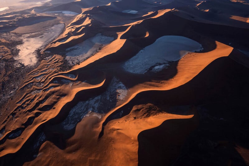Aerial view of sand dunes in a desert at sunrise or sunset, with long shadows and patches of white salt or dry clay contrasting against the dark sand ridges.