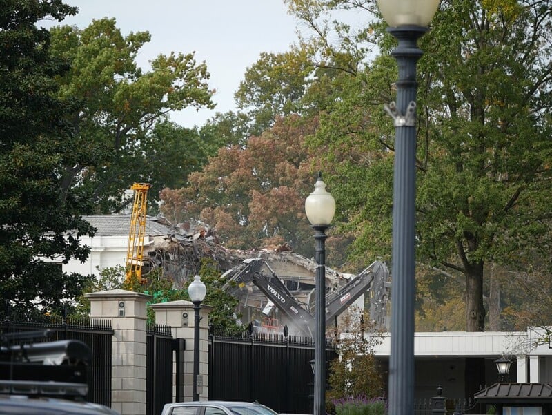 A demolition excavator tears down part of a large building behind a gated fence, surrounded by trees and street lamps, with debris and dust visible in the background.