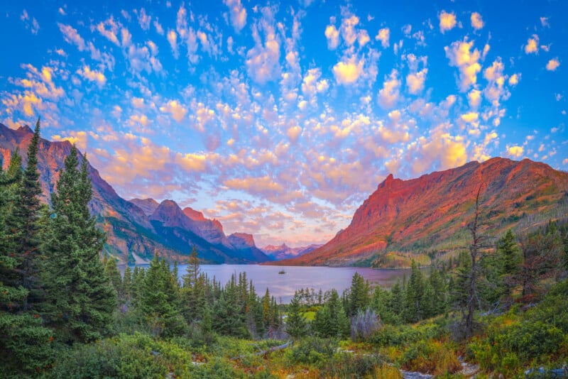 A vibrant sunrise casts warm light on rugged mountains and a lake surrounded by pine trees, with a sky filled with scattered, colorful clouds. The scene is lush and tranquil, showcasing natural beauty and vivid colors.