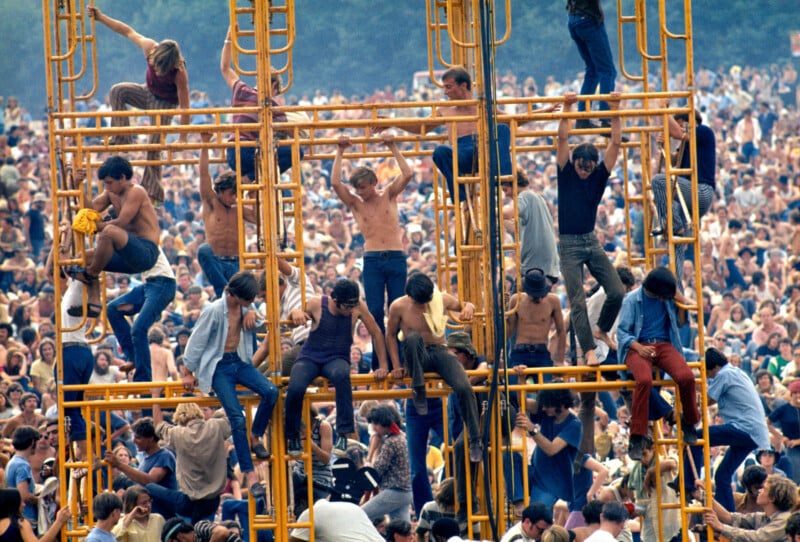 A large crowd gathers outdoors as many shirtless young people climb on yellow scaffolding, overlooking the masses below, creating a vibrant, energetic scene at a music festival.