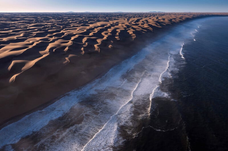 Aerial view of a vast desert with rippling sand dunes meeting the ocean, where waves crash along the shoreline under a clear blue sky.