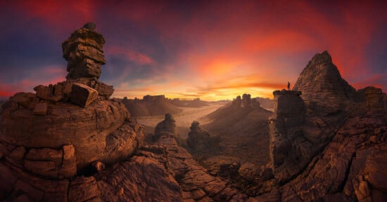 A person stands on a jagged rock formation, overlooking a dramatic, rocky desert landscape at sunset, with vivid orange and purple clouds filling the sky.