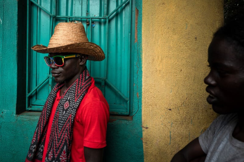 Un hombre con sombrero de paja, gafas de sol de colores, una camisa roja y una bufanda estampada se encuentra frente a una ventana verde azulado y una pared amarilla, mientras que una mujer con una camisa gris se encuentra en las sombras a la derecha.