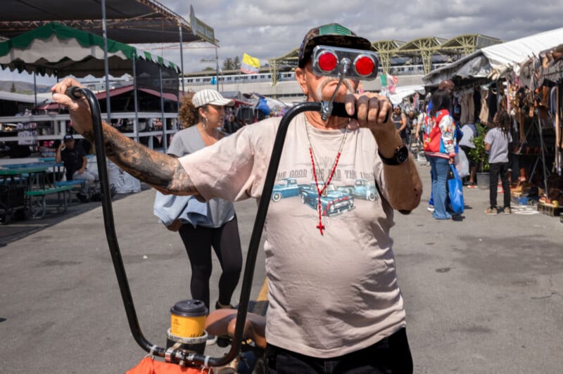 A man with tattooed arms wearing unique red goggles and a cap rides a bicycle at an outdoor market, with other people and vendor stalls visible in the background under a partly cloudy sky.