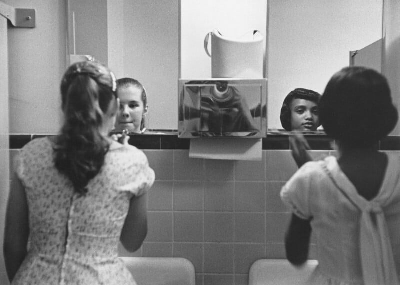 Two girls in dresses stand at sinks in front of a bathroom mirror, with their reflections visible. Another girl’s reflection, partially obscured, appears between them. The scene is in black and white.