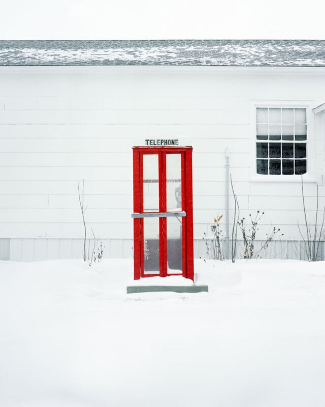 Una cabina telefónica de color rojo brillante se alza sobre la espesa nieve, frente a un edificio blanco con ventanas, en marcado contraste con la sencilla escena invernal.