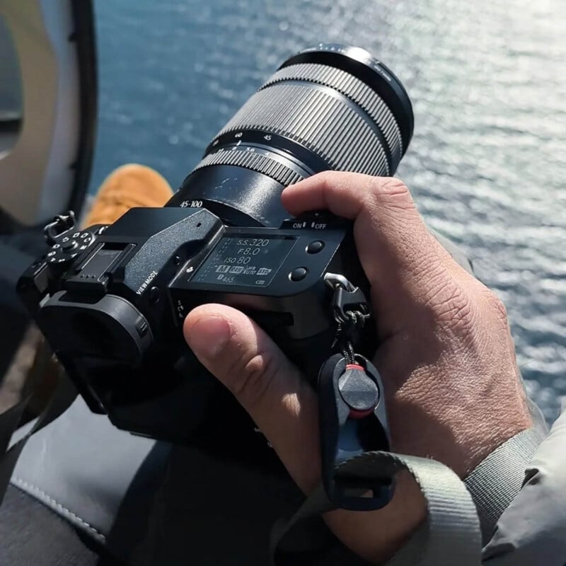 A person holds a digital camera with a large zoom lens, sitting near an open window or edge, with water visible below in the background.