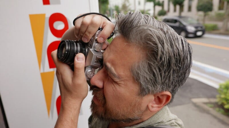 Un hombre barbudo y de pelo gris se acerca una cámara a los ojos y toma una fotografía al aire libre cerca de un cartel blanco y rojo, con la calle y los coches aparcados visibles al fondo.