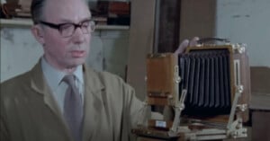 A man wearing glasses and a beige coat stands beside a large, vintage wooden camera with a bellows, inside a workshop or studio with shelves in the background.