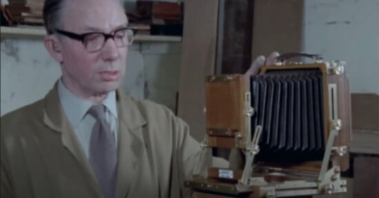 A man wearing glasses and a beige coat stands beside a large, vintage wooden camera with a bellows, inside a workshop or studio with shelves in the background.
