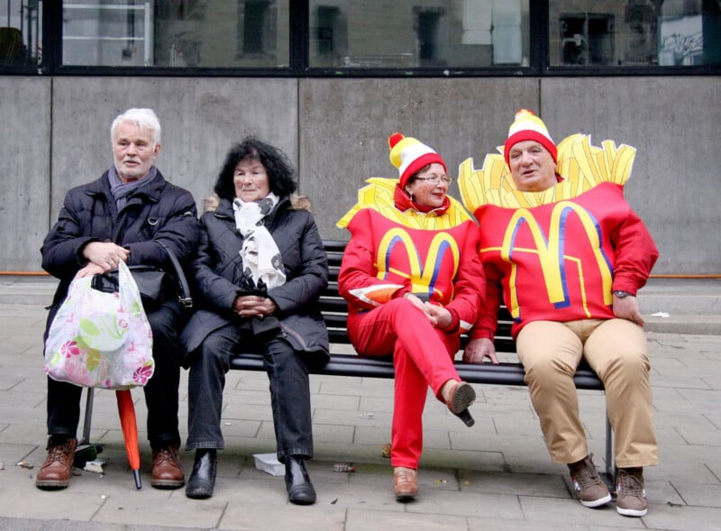 Four adults sit on a bench; two are dressed in regular winter clothing, while the other two wear bright costumes resembling McDonald's French fries. All face forward, in an urban setting.