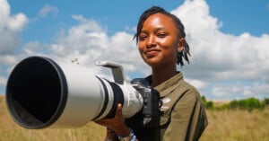 A woman standing outdoors in a grassy field holds a large professional camera with a long telephoto lens, smiling at the camera. The sky is blue with scattered clouds.