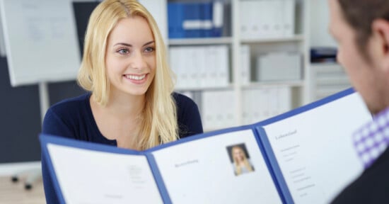 A woman with long blonde hair smiles while sitting across a desk from a person holding a folder with her resume and photo during a job interview in an office setting.