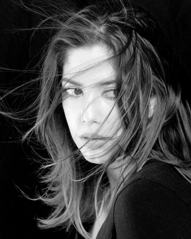 Black and white portrait of a woman with long hair blowing across her face, gazing directly at the camera against a dark background.