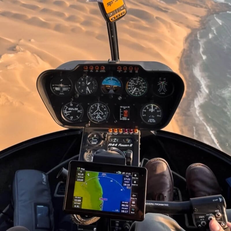 View from inside a helicopter cockpit flying over desert dunes and coastline, showing various navigation instruments and screens, with the pilot's feet visible on the controls.