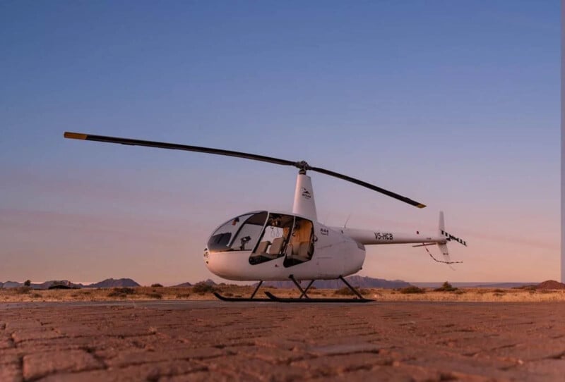 A white helicopter is parked on a cracked, dry ground at sunset, with mountains visible in the distant background under a clear sky.