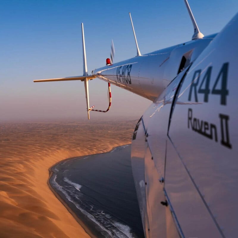 Aerial view from a helicopter flying along a coastline, with sandy desert dunes meeting the ocean. The side of the helicopter displays "R44 Raven II." The sky is clear and the scene is bathed in warm light.