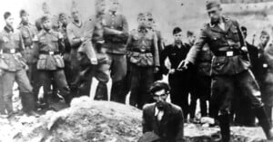 A group of uniformed soldiers stand outdoors as one soldier points a gun at a kneeling man with glasses, suggesting an execution scene; other men observe somberly in the background. The image is in black and white.