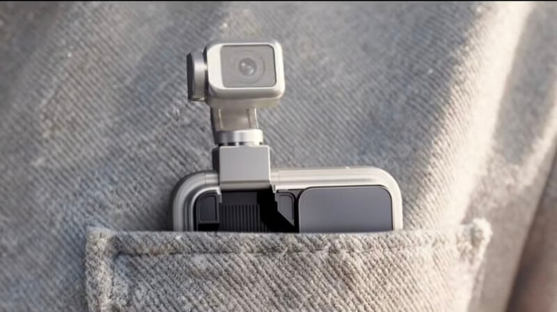 A compact camera with a gimbal is attached to a smartphone and tucked into the chest pocket of a light gray textured jacket.