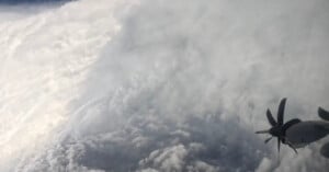 Aerial view from an aircraft flying near the edge of a dense, swirling hurricane, with the aircraft’s propeller visible on the right side and thick clouds dominating the scene.