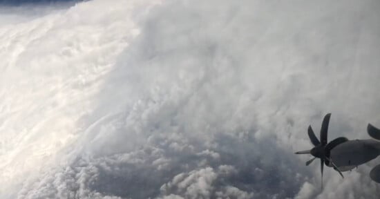 Aerial view from an aircraft flying near the edge of a dense, swirling hurricane, with the aircraft’s propeller visible on the right side and thick clouds dominating the scene.