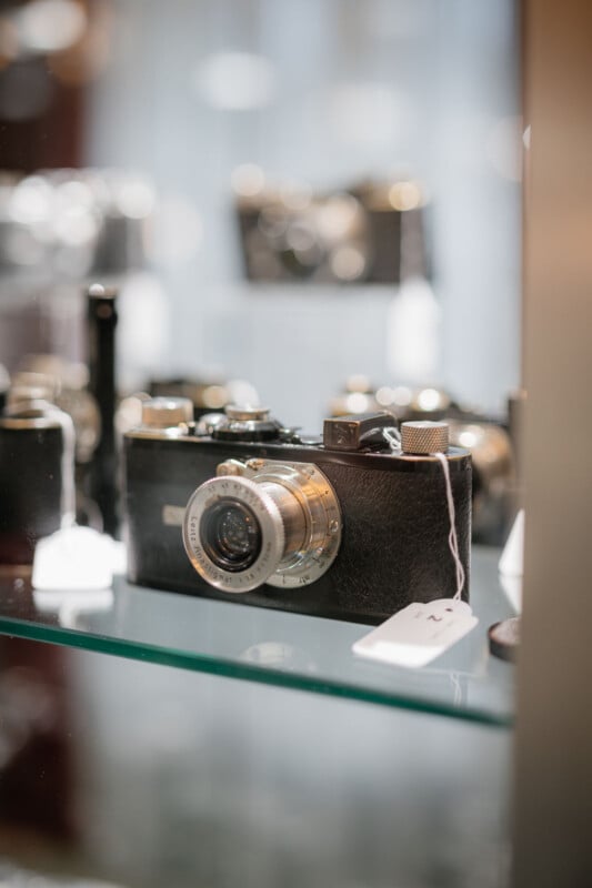 A vintage black and silver camera with a textured body sits on a glass shelf, accompanied by a white price tag. Other old cameras are blurred in the background.