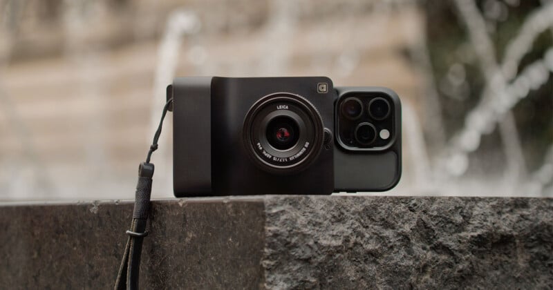 A smartphone with a Leica camera attachment and wrist strap rests on a stone surface, with a blurred fountain in the background.