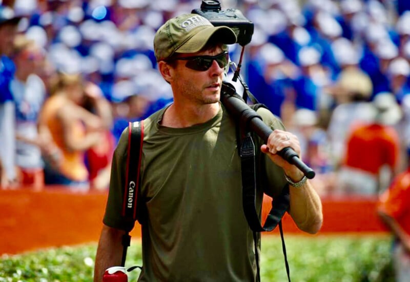 A man wearing sunglasses and a green shirt carries a large video camera on his shoulder in an outdoor, crowded event. He has a Canon camera strap over his shoulder and is surrounded by people in the background.