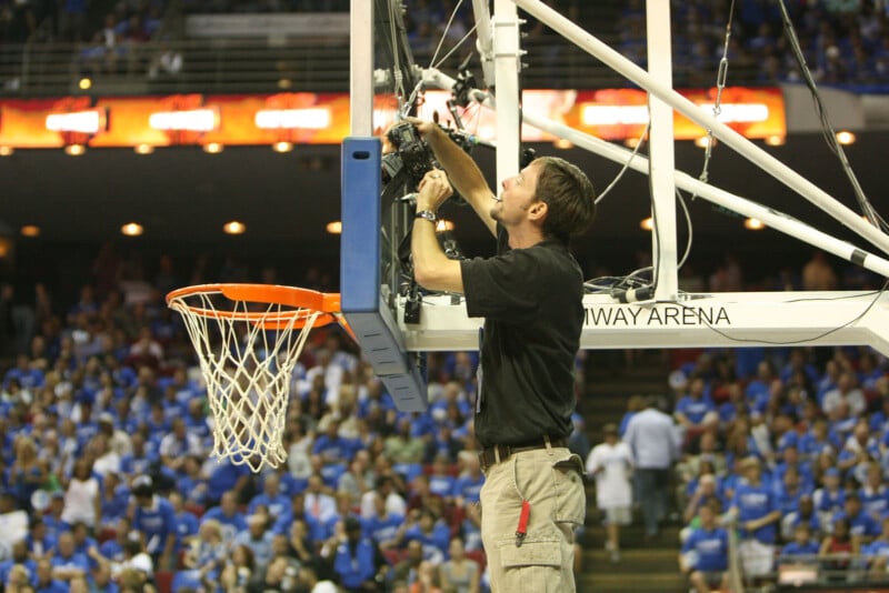 A technician stands on a platform adjusting a camera mounted on a basketball hoop's backboard in a crowded arena filled with fans wearing blue shirts.