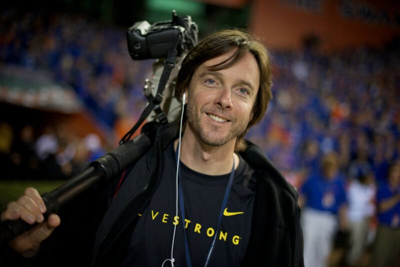 A smiling man with shoulder-length brown hair holds a video camera on his shoulder. He wears a black "LIVESTRONG" shirt and an earbud, standing in a crowded stadium with blurred spectators in the background.