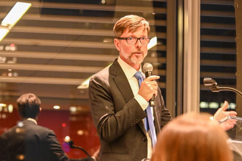A man in a suit and glasses speaks into a microphone at an indoor event, standing near a window with blinds and city lights visible outside. Another person is partially visible in the foreground.