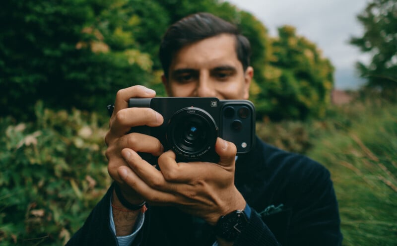 A man outdoors, holding a camera with a smartphone attached to it, aims the lens toward the viewer. Green foliage is blurred in the background.