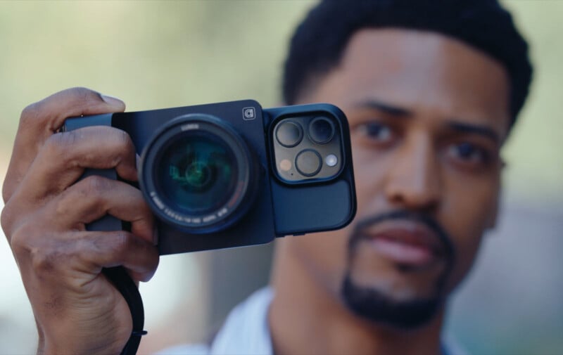 A man holds a camera with a smartphone attached to the back, combining both devices. The focus is on the camera, while the man’s face is slightly blurred in the background.