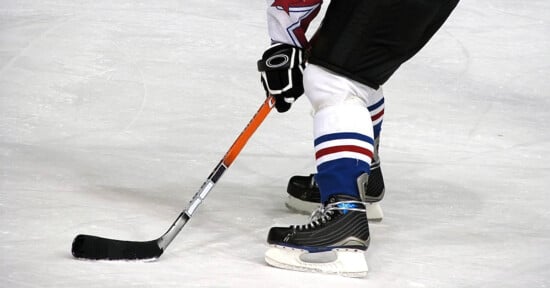 A hockey player on ice in black skates and blue-and-white socks controls a puck with a black stick, preparing to make a move during a game.