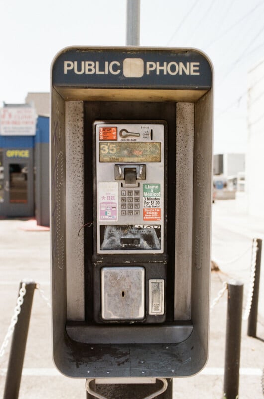 A weathered public payphone stands outdoors, displaying faded stickers and instructions. The phone booth is empty, with a coin slot and receiver cradle visible. Buildings and a parking lot are in the background.
