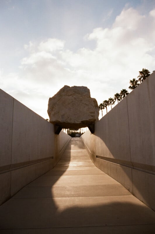 A large boulder sits elevated above a concrete walkway flanked by tall walls, with palm trees and a partly cloudy sky in the background.